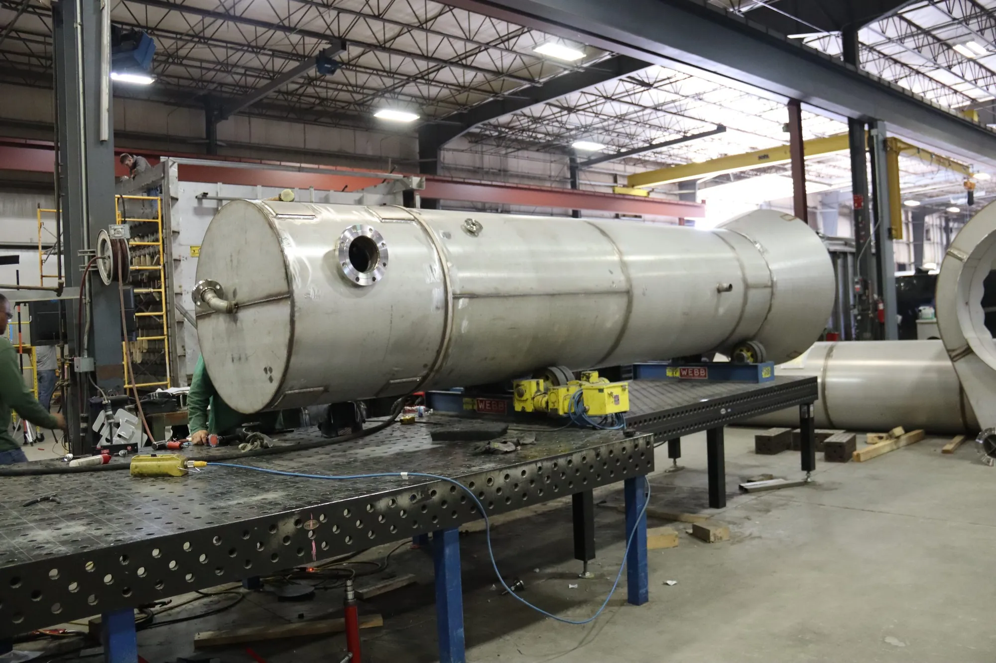 Large horizontal stainless steel process vessel with nozzles staged on turning stands beside a welding table at Northern Manufacturing