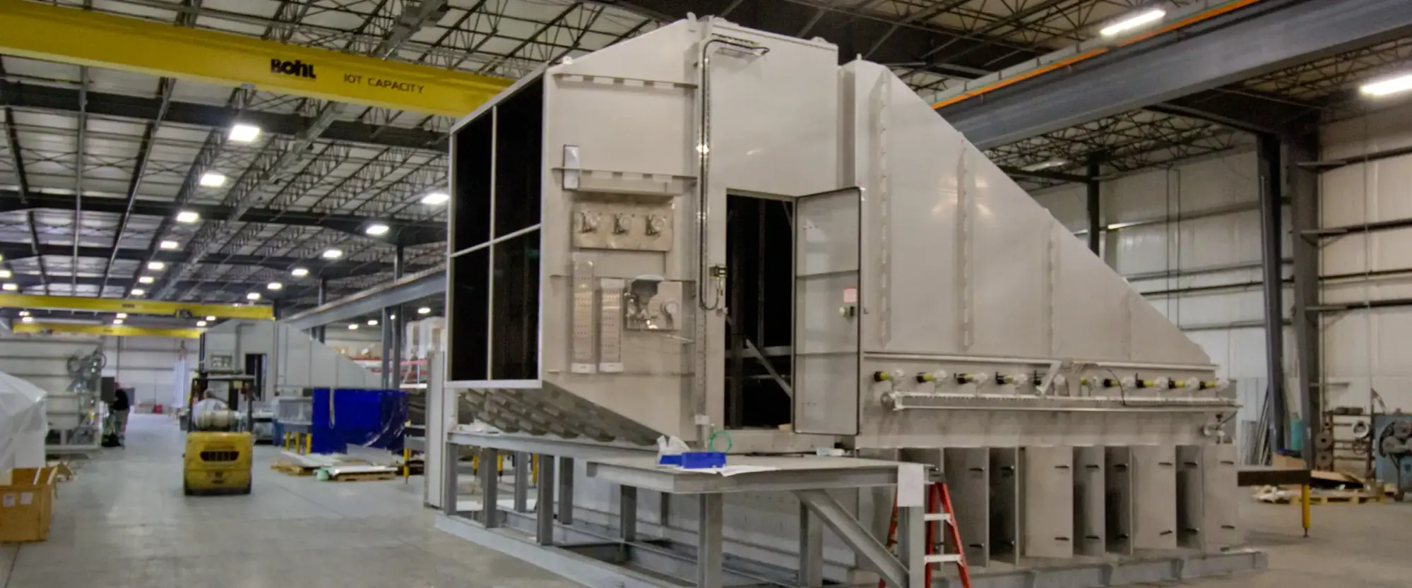 Very large fabricated enclosure with a sloped top and access door under construction in the Northern Manufacturing bay, with a forklift alongside