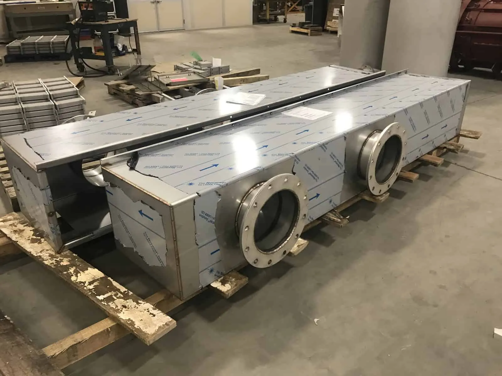 Two long rectangular stainless duct sections with bolted flange ports and protective film, staged on pallets in the Northern Manufacturing shop