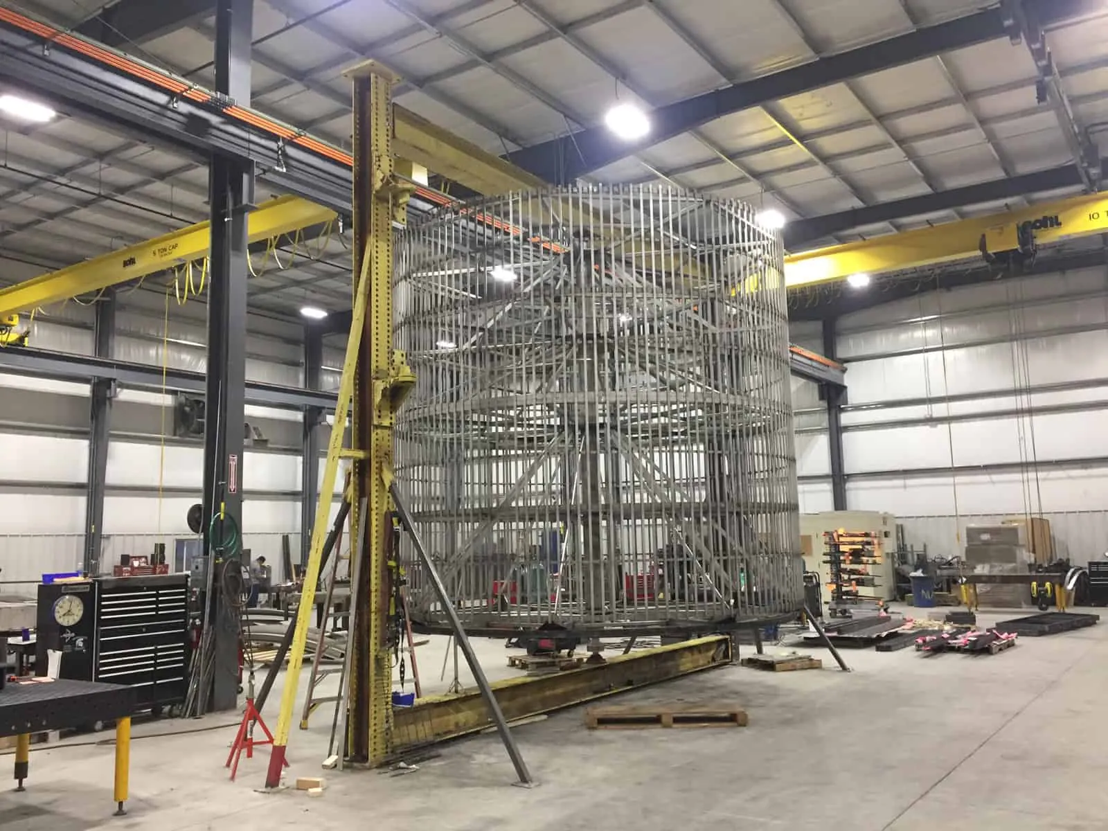Large cylindrical stainless steel frame assembly fixtured for welding under overhead cranes